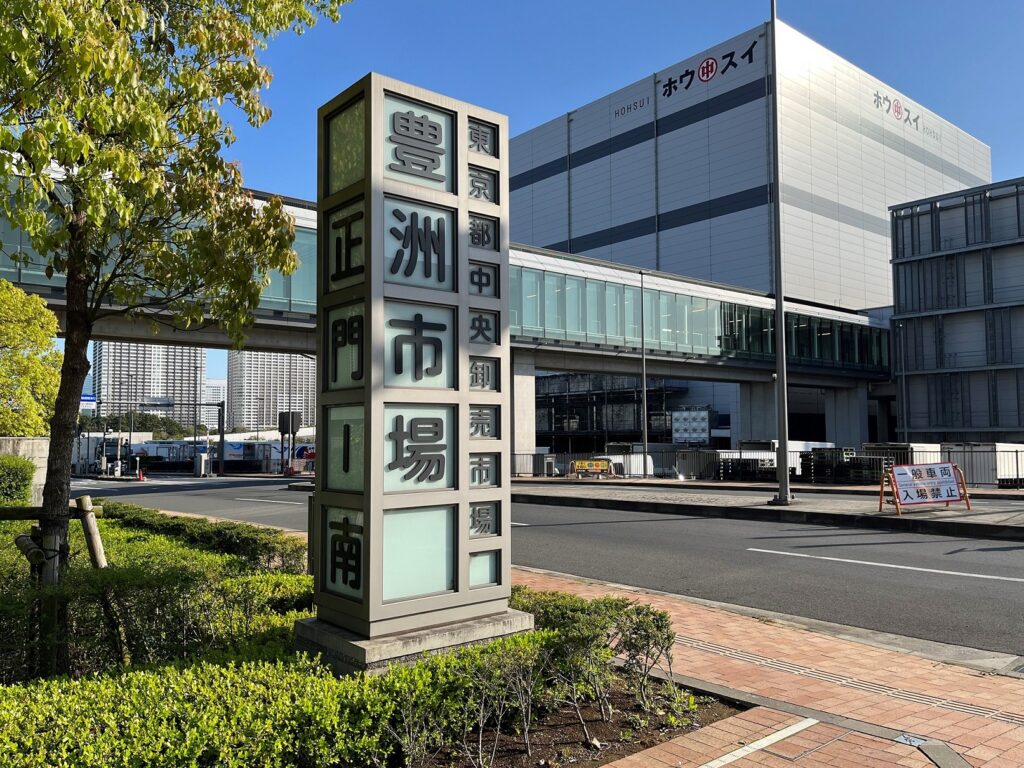 Un panneau vertical indique en japonais "Marché de Toyosu". En arrière-plan, les infrastructures du marché au poisson