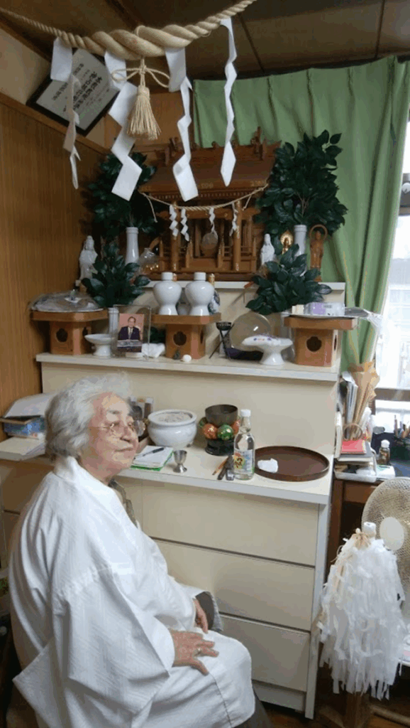 Une femme âgée habillée de blanc regarde de profil et se recueille, agenouillée, devant un autel chargé d'objets dévotionnels.