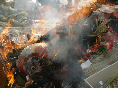 Des flammes lèchent un daruma en train de brûler dans les feux du Dondoyaki. A côté de lui, on peut voir des décorations du Nouvel An en train de brûler elles aussi. 