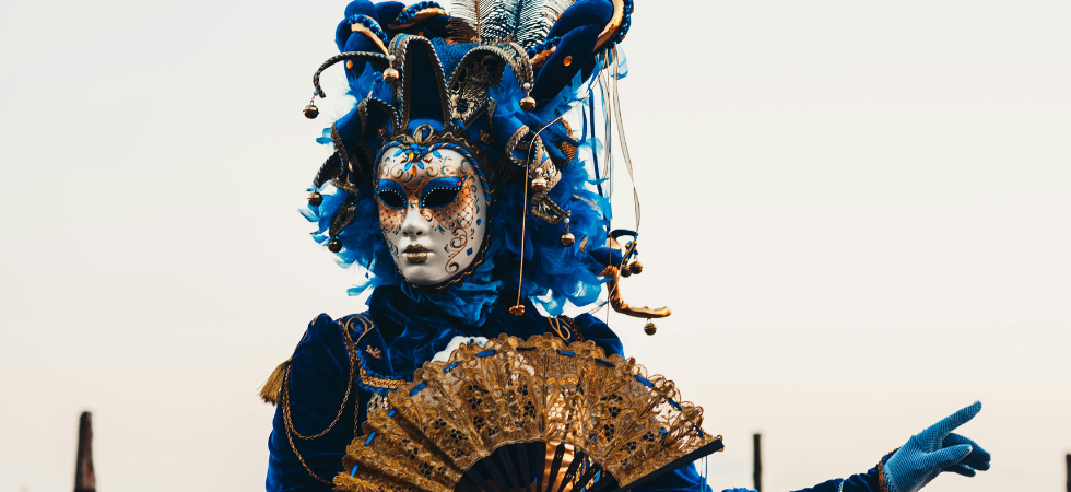 Photo d'une personne portant un costume au carnaval de Venise. Son cotsume est dans les ton bleus et dorés, elle porte un masque caractéristique de cette fête.