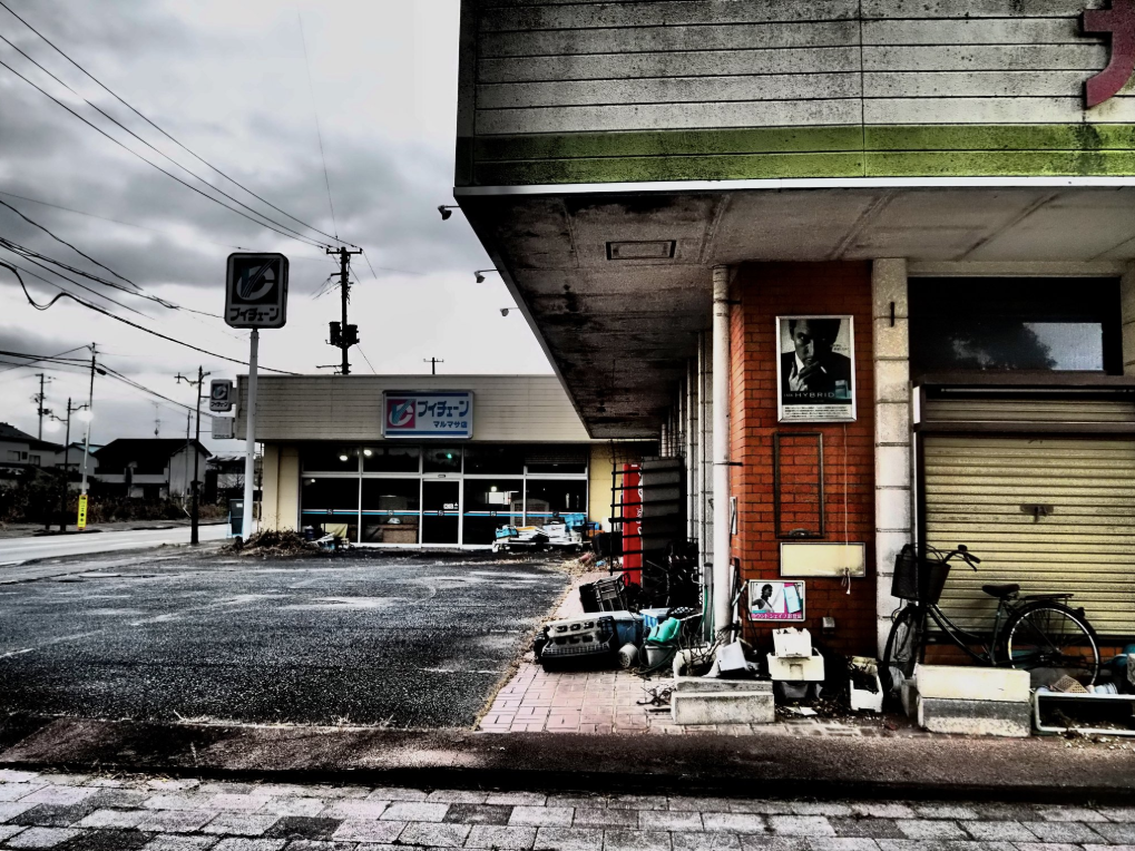 Au premier plan, un bâtiment abandonné avec un vélo et des objets éparpillés sur le sol. En arrière-plan à gauche, un parking désert.