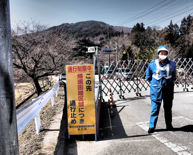 A droite, une femme en tenue de travail monte la garde devant un barrage routier. A gauche, un panneau jaune vertical indique en japonais que la route mène vers une zone évacuée.