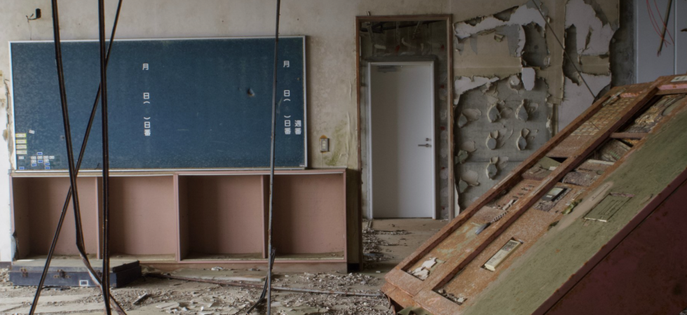 Intérieur d'une salle de classe abandonnée. Au centre, un tableau noir. A droite, une bibliothèque effondrée