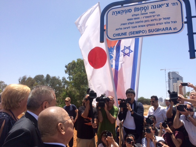 Scène de foule. A droite, des photographes immortalisent des visiteurs pour célébrer l'ouverture d'une rue au nom de Chiune Sugihara. En arrière-plan, des drapeaux japonais et israélien ainsi qu'un panneau en hébreu.