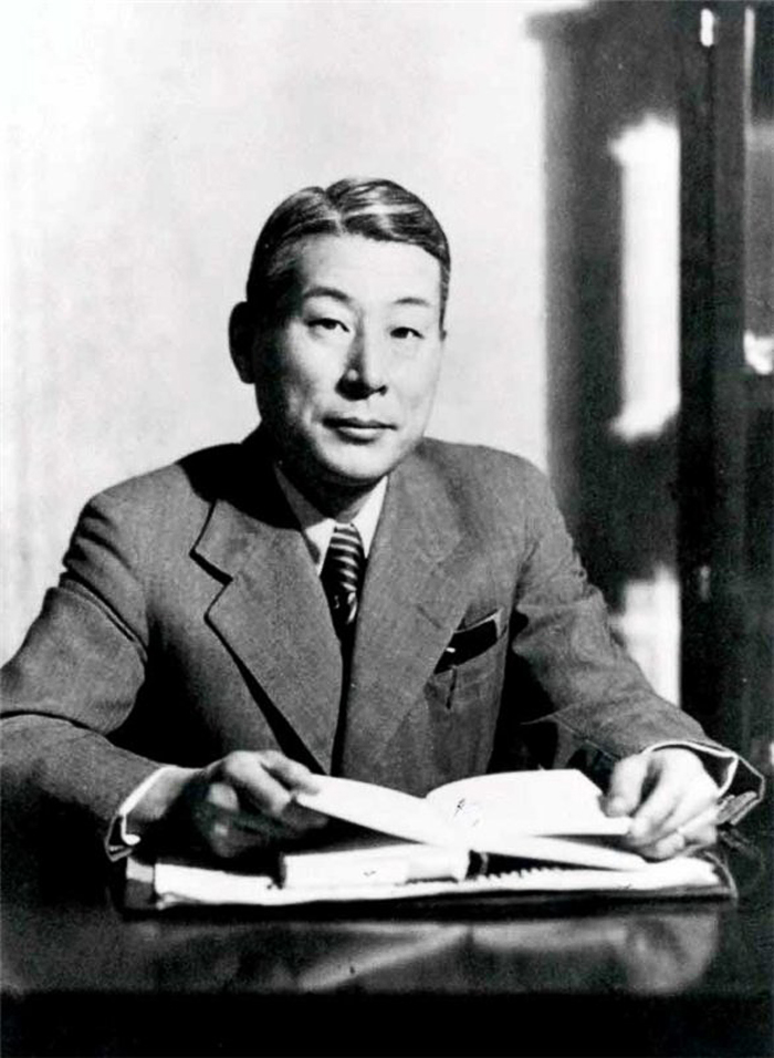 Portrait de Chiune Sugihara en noir et blanc, de face et assis à un bureau, tenant un livre ouvert.